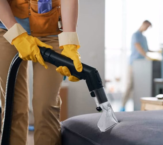 A woman wearing yellow gloves is cleaning a couch with a cloth, focusing on removing dirt and stains